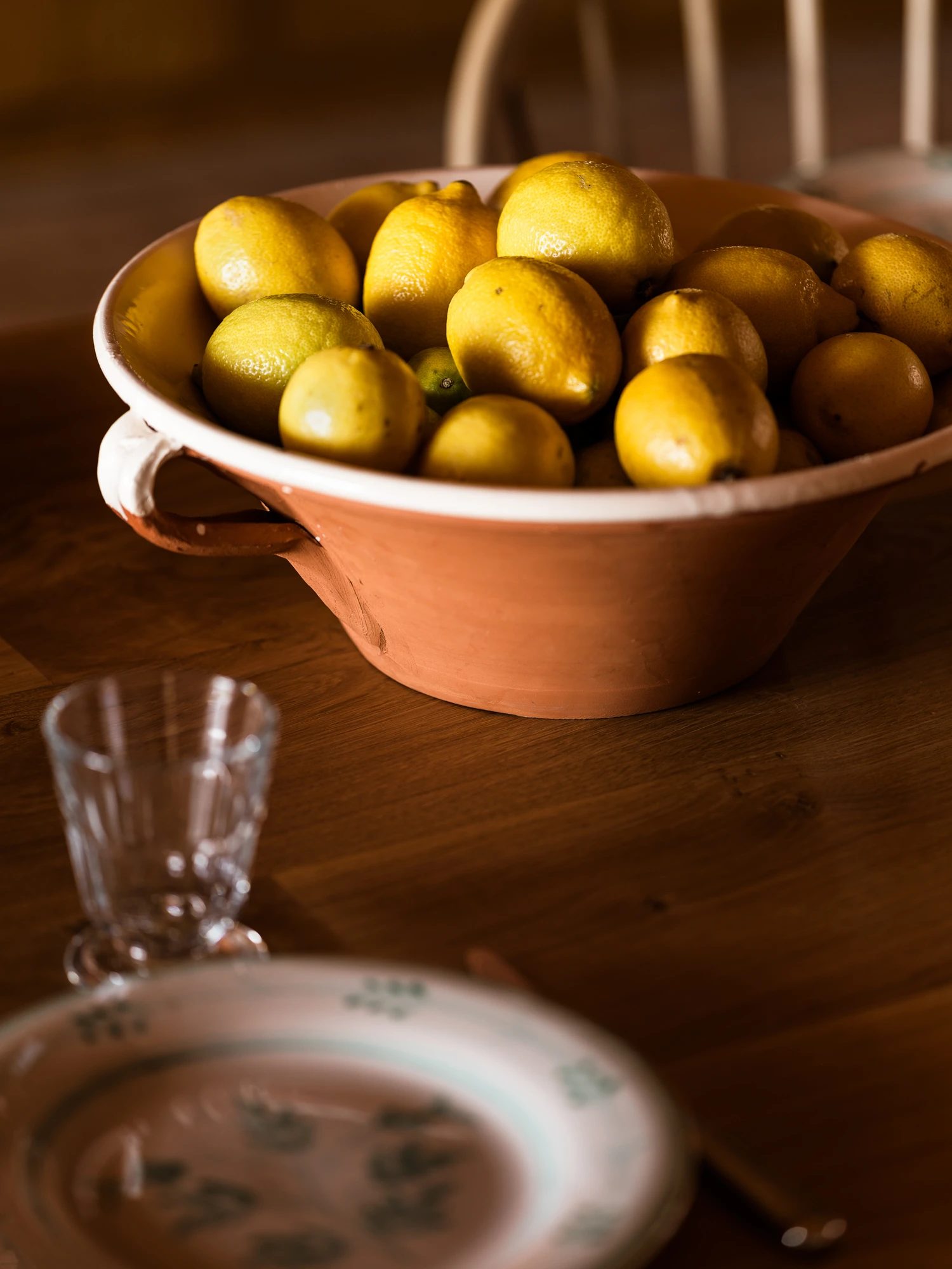 A bowl of lemons, cut glass and a painted plate on a wooden table.