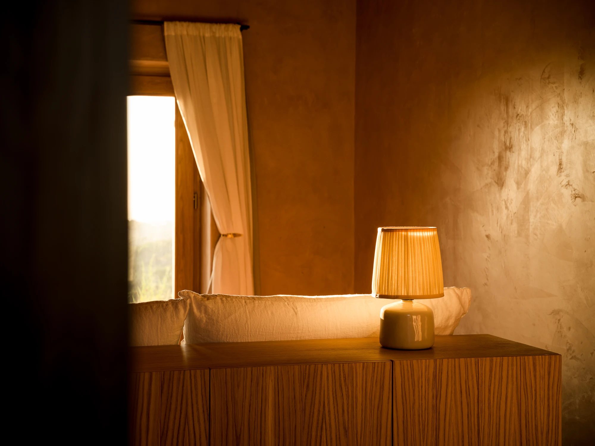 A bedroom corner at dusk: a low bed beneath a curtained window, lit by a pleated lamp.