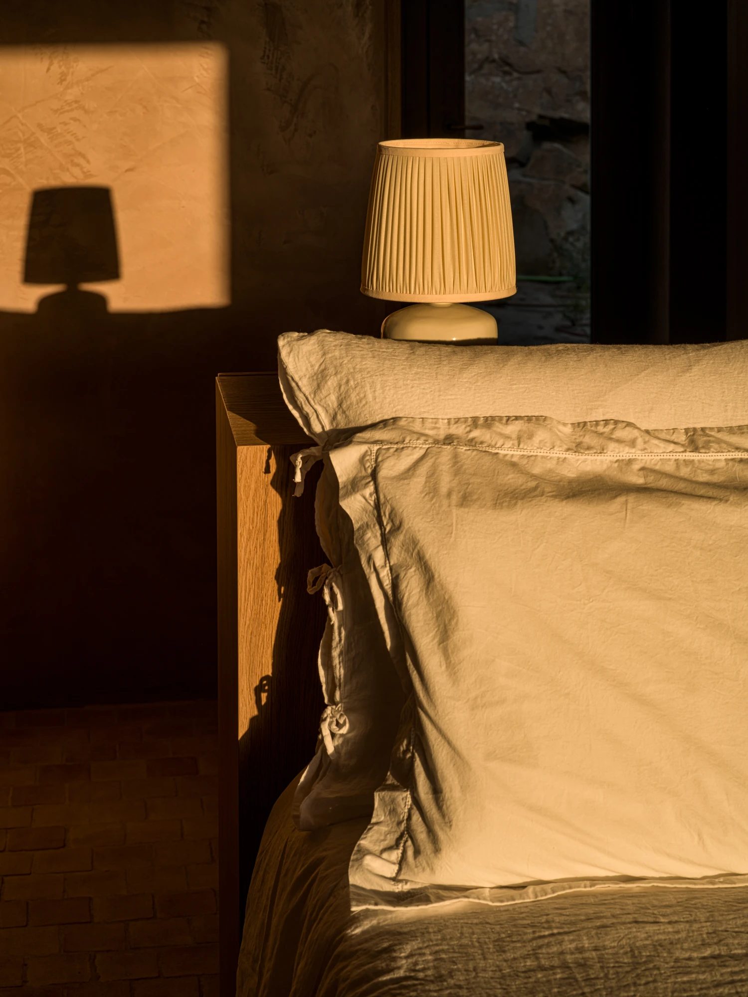 A bed dressed in linen, with morning light falling across the rammed-earth wall.